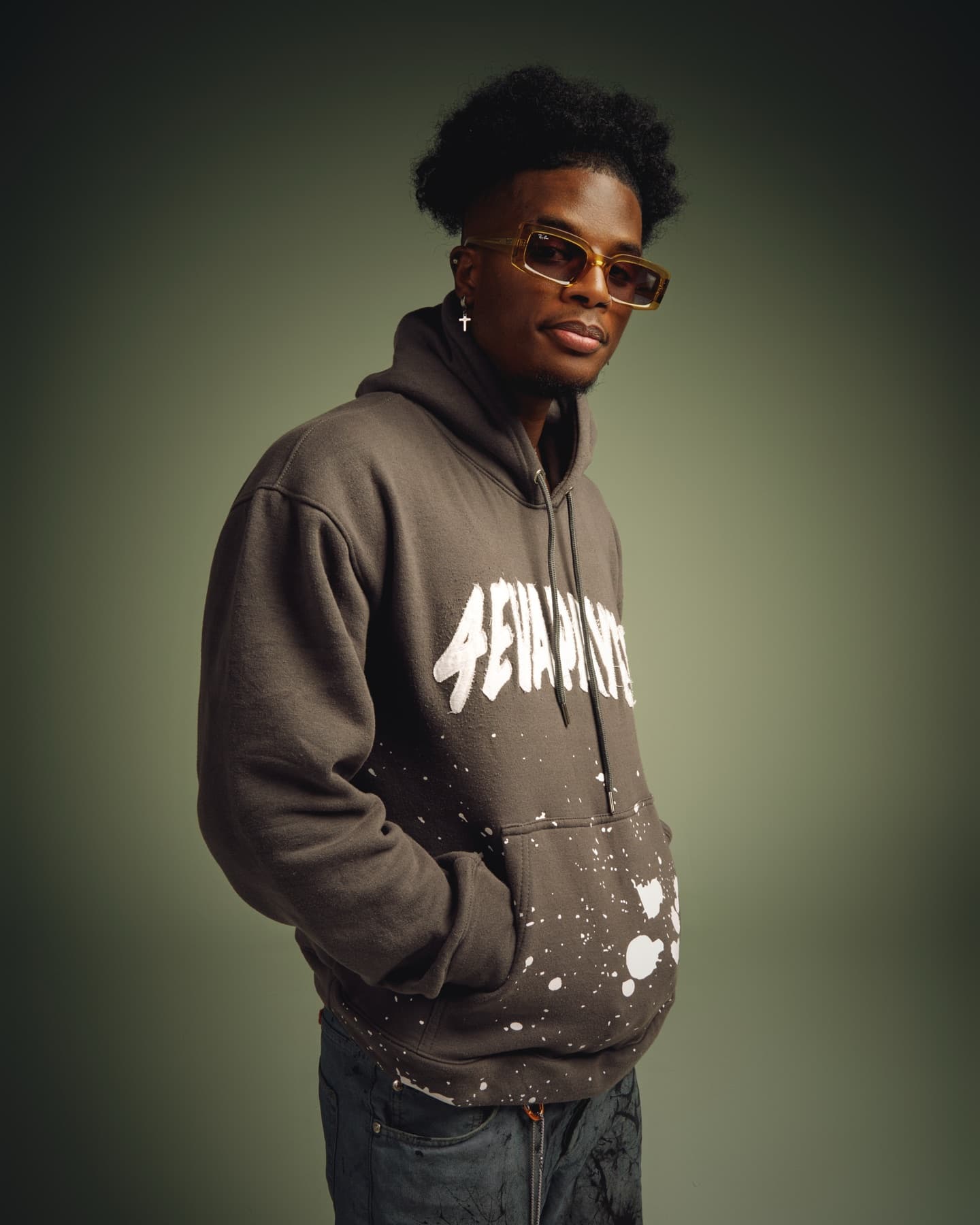 Model in sunglasses and a paint-splattered hoodie against an olive backdrop